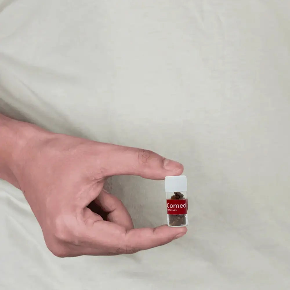 A hand holding a small clear plastic vial containing dark Vastu remedy crystals.
